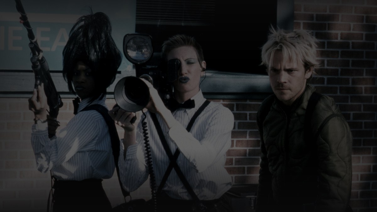 Still from Cecil B. Demented (2000) — tmdb backdrop (key fKHf0o9JQ62Ak06CUyqvJKbu4v0); genuine production still, the gang with honey whitlock at the brick-wall holdout. source luma 113.46, pre-darkened 0.78, baked at factor 0.45, lanczos to 1200x675, on-disk luma 30.24