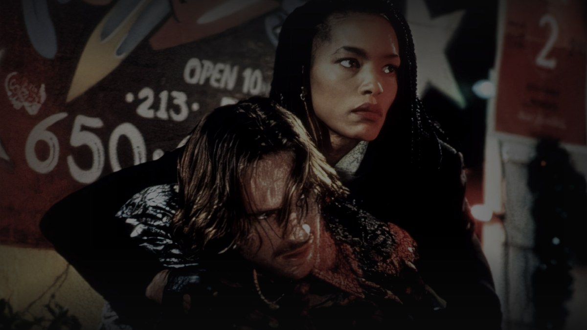 Still from Strange Days (1995) — letterboxd og:image (1200x675 crop); genuine production still of mace shielding lenny against the la graffiti backdrop. source luma 76.78, pre-darkened 1.0, baked at factor 0.45, lanczos to 1200x675, on-disk luma 26.74